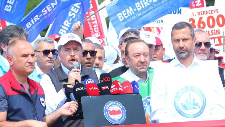Mikrofonla konuşan bir kişi, çevresindeki kalabalık ve sendika bayrakları önünde kürsüde duruyor.