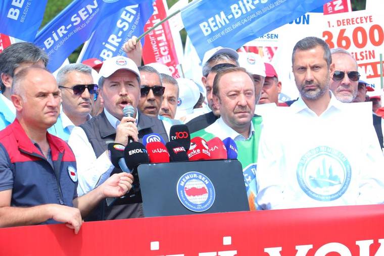 Mikrofonla konuşan bir kişi, çevresindeki kalabalık ve sendika bayrakları önünde kürsüde duruyor.