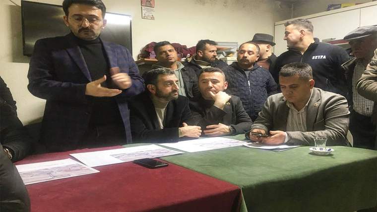 Kapalı bir odada, masanın çevresinde oturan ve ayakta duran bir grup erkek görülüyor.