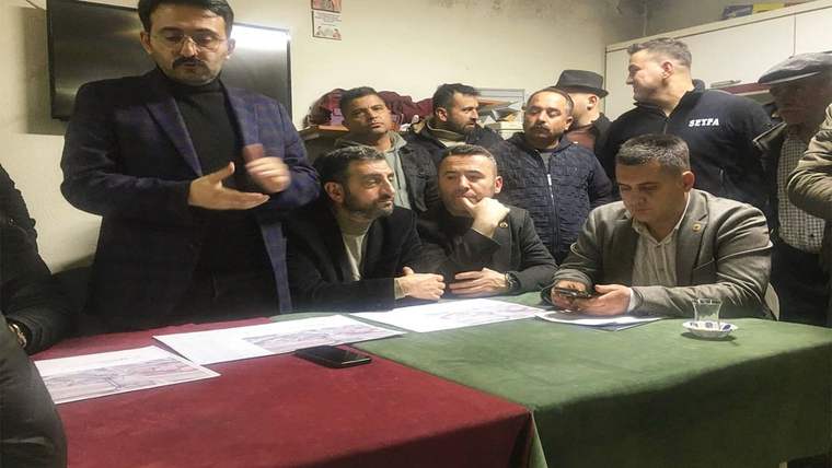 Bir odada, masa çevresinde toplanan erkekler; masada belgeler, telefonlar ve küçük bir Türk bayrağı bulunuyor.