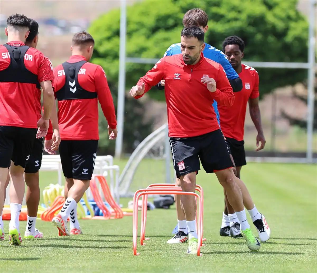Antrenman sahasında, kırmızı forma giymiş futbolcular, engellerin etrafında koşuyor.