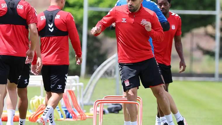 Antrenman sahasında, kırmızı forma giymiş futbolcular, engellerin etrafında koşuyor.