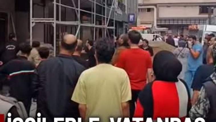 Bir grup insan, inşaat alanında toplanmış, bazıları tartışıyor. Arka planda binalar ve bir dükkan görünmekte.