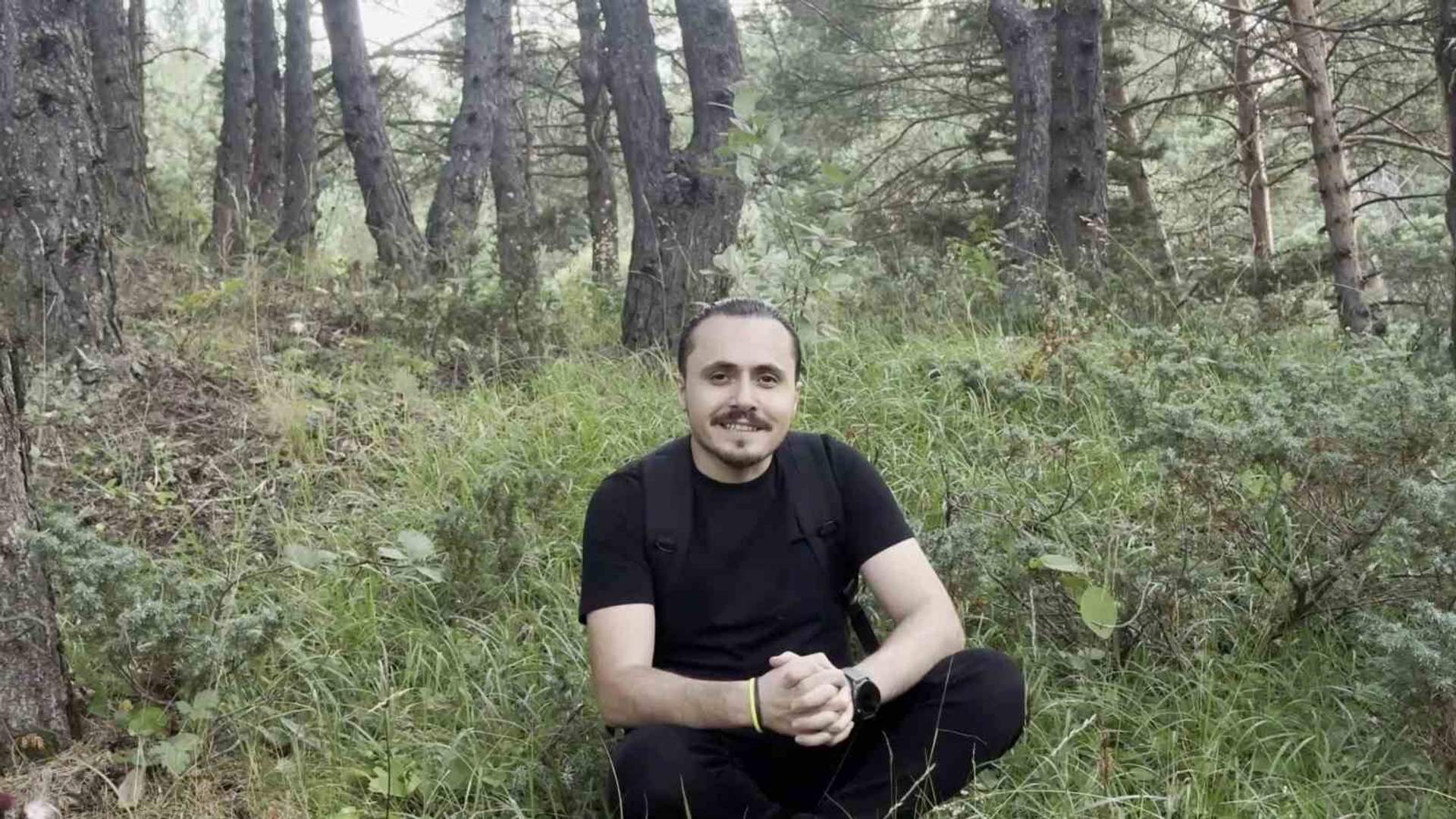Ormanlık alanda oturan siyah tişörtlü bir adam, ellerini birleştirerek kameraya bakıyor.