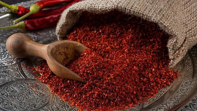 Çuvaldan işlemeli metal tepsiye dökülen pul biberin yanında tahta kaşık ve kırmızı biberler görülüyor.