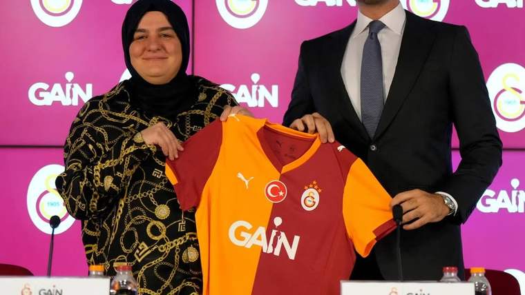 İki kişi, üzerinde Galatasaray arması ve GAİN logosu bulunan turuncu-kırmızı formayı tutuyor.