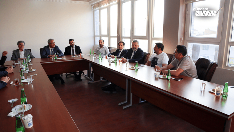 Bir toplantı odasında, uzun bir masanın etrafında oturan yedi kişi, su ve içecek şişeleriyle birlikte görüntüleniyor.