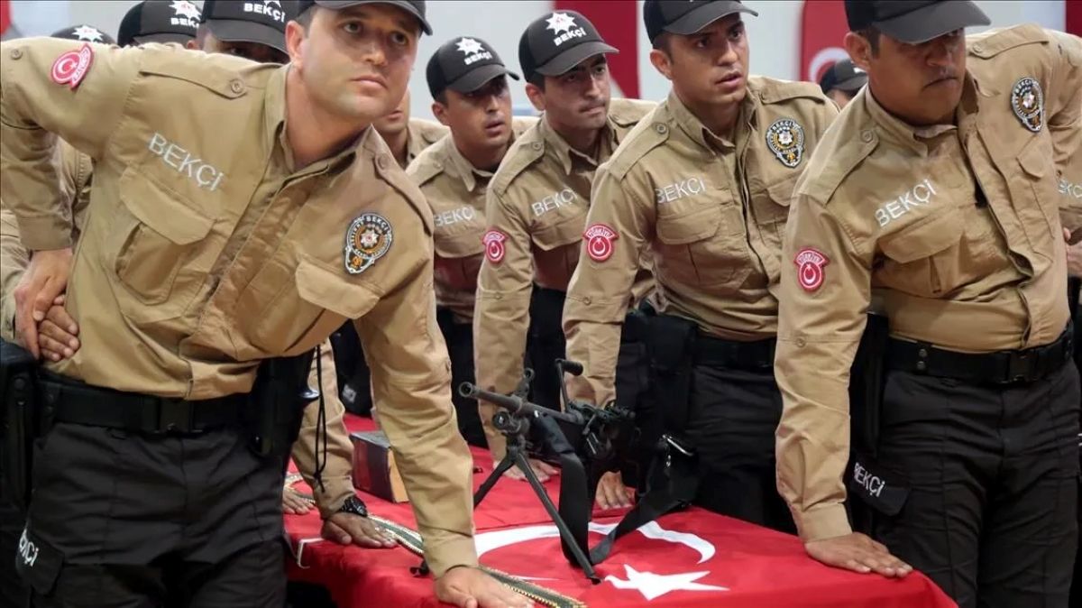 Üzerlerinde “BEKÇİ” yazısı bulunan üniformalı kişiler, Türk bayrağı örtülü bir yüzeyin yanında duruyor.