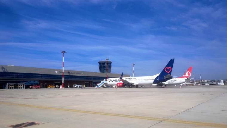 Apronda park etmiş iki yolcu uçağı, terminal binası ve açık hava altında geniş beton zemin görülüyor.