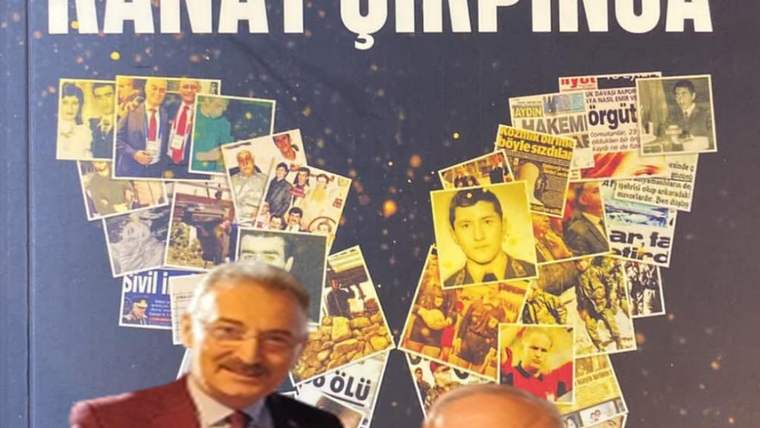 İki kişi, arka planda çeşitli fotoğrafların yer aldığı bir duvarın önünde duruyor.