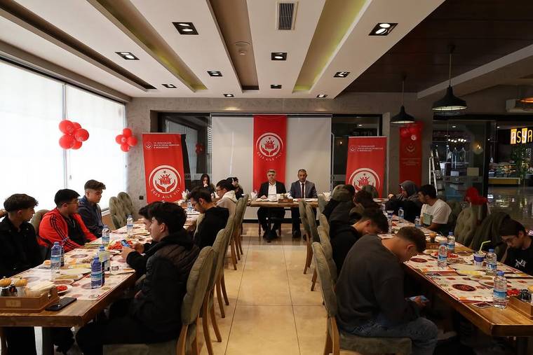 Restoran salonunda uzun masalarda oturan öğrenciler, karşılarındaki masada konuşan iki kişiyle birlikte görülüyor.