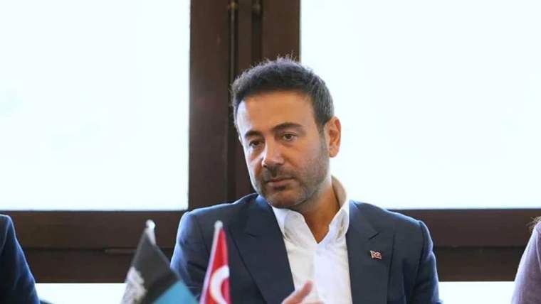 Koyu renk takım elbiseli bir kişi, önündeki Türk bayrağı ve başka bir bayrakla birlikte oturuyor.