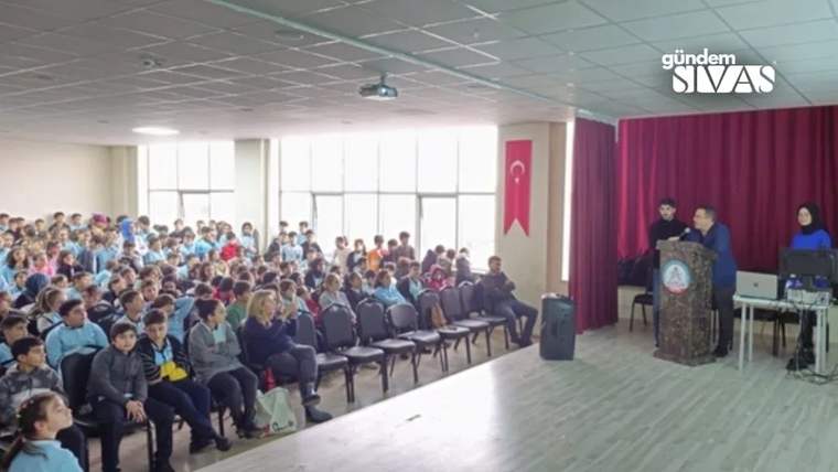 Salonda oturan öğrenciler, Türk bayrağı ve perde önündeki kürsüde duran iki kişi görülüyor.