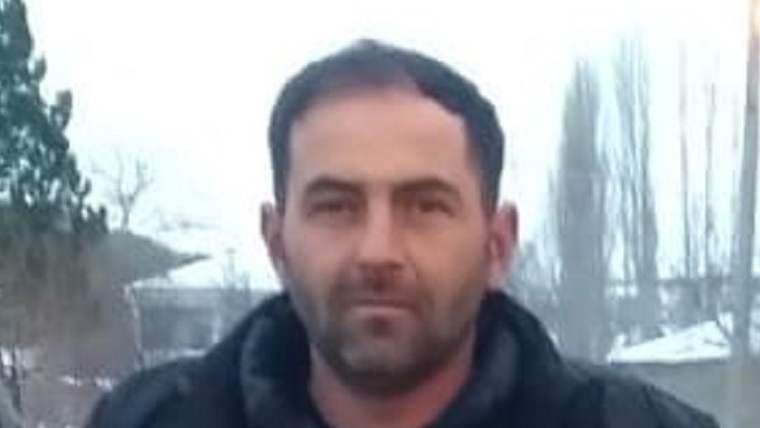 Siyah montlu, sakallı bir adam dış mekânda kameraya doğru bakıyor; arka planda ağaçlar ve yapılar görülüyor.