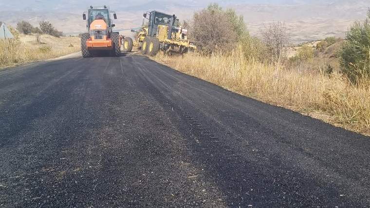 Kırsal alanda siyah asfaltla kaplanan yolda turuncu silindir ve sarı greyder iş makinesi bulunuyor.