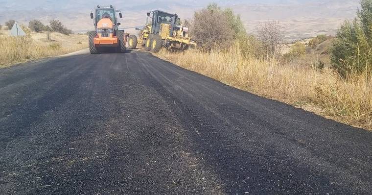 Kırsal alanda siyah asfaltla kaplanan yolda turuncu silindir ve sarı greyder iş makinesi bulunuyor.