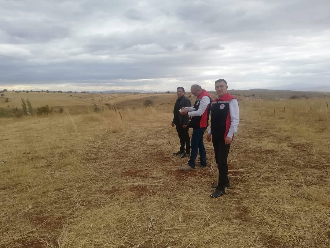 Sarı otlarla kaplı geniş bir arazide, kırmızı-siyah yelekli üç kişi yan yana duruyor.