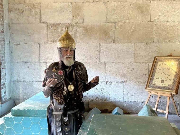 Taç ve zırh benzeri kıyafet giyen sakallı kişi, taş duvar önünde duruyor; yanında yazılı çerçeve var.