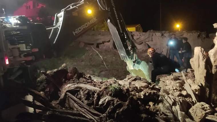 Gece ışıkları altında bir kepçe, yıkıntılar arasındaki enkazı kaldırırken çevrede insanlar görülüyor.