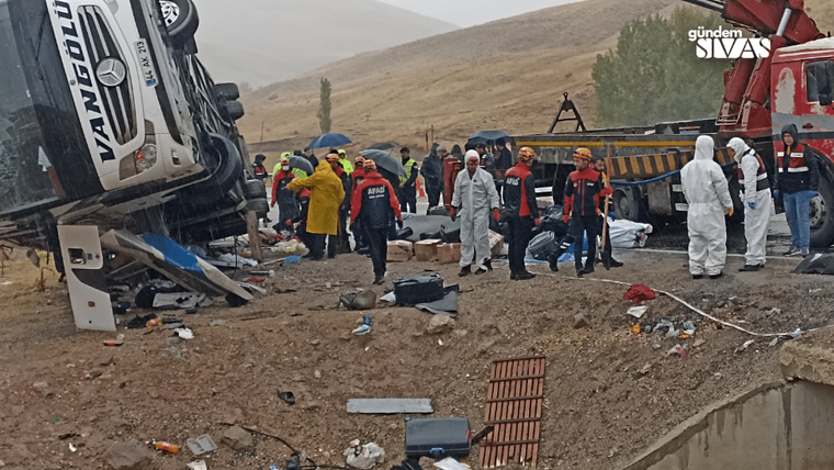 Devrilmiş bir otobüsün yanında yağmur altında insanlar ve ekipler, enkaz parçaları arasında duruyor.