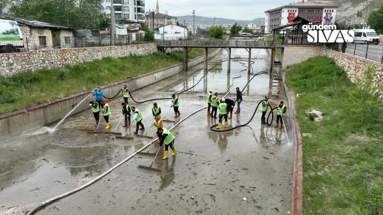 Reflektörlü yelek giyen kişiler, beton kanaldaki su ve çamuru hortumlarla temizliyor.
