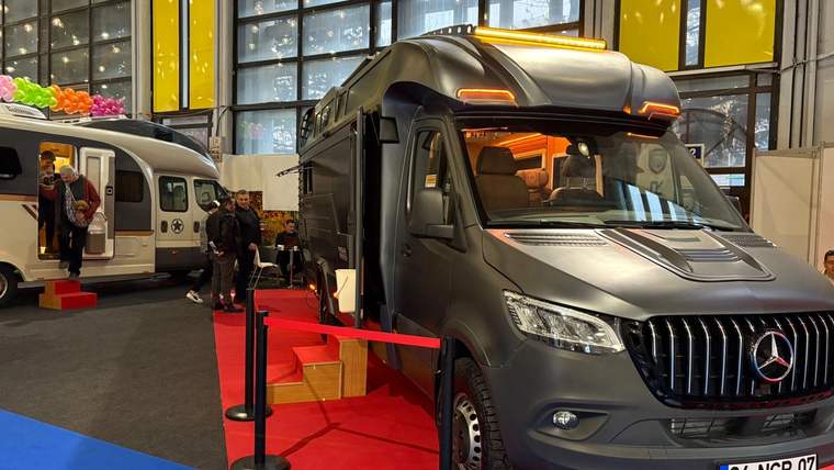Fuar salonunda kırmızı şeritlerle çevrili büyük bir karavan ve yanında sergilenen başka bir karavan görülüyor.