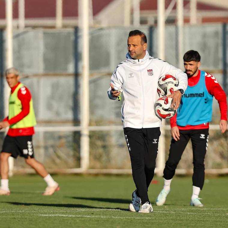 Antrenman sahasında üç futbolcu yürürken, ortadaki kişi kucağında iki futbol topu taşıyor.