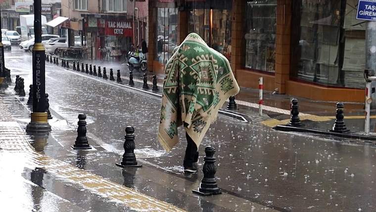 Dolu yağışı altındaki caddede, desenli battaniyeyle başını örten bir kişi yürürken görülüyor.