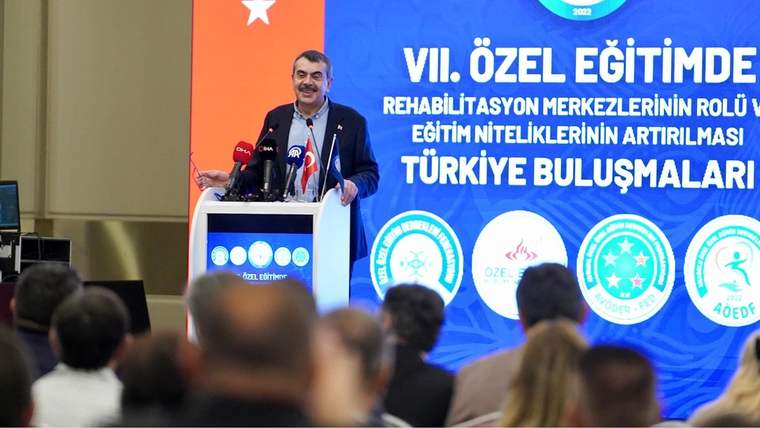 Kürsüde konuşan bir kişi, önündeki dinleyicilere hitap ediyor; arkada özel eğitim buluşmalarını anlatan pankart bulunuyor.