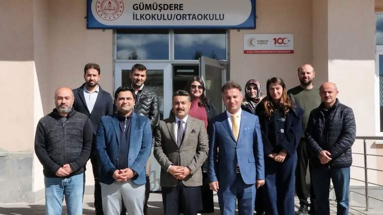 Gümüşdere İlkokulu/Ortaokulu tabelası önünde, aralarında takım elbiseli kişilerin de bulunduğu dokuz kişi duruyor.