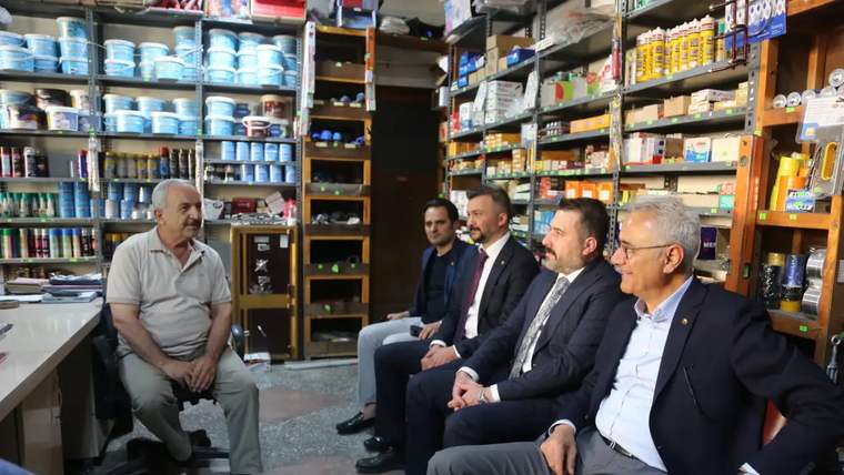 Bir dükkânda raflardaki ürünlerin önünde oturan beş erkek, karşılarındaki esnafı dinliyor.