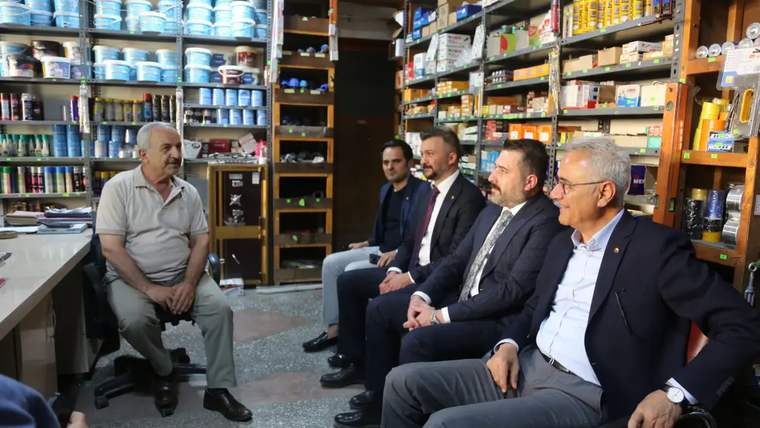 Bir dükkânda raflardaki ürünlerin önünde oturan beş erkek, karşılarındaki esnafı dinliyor.