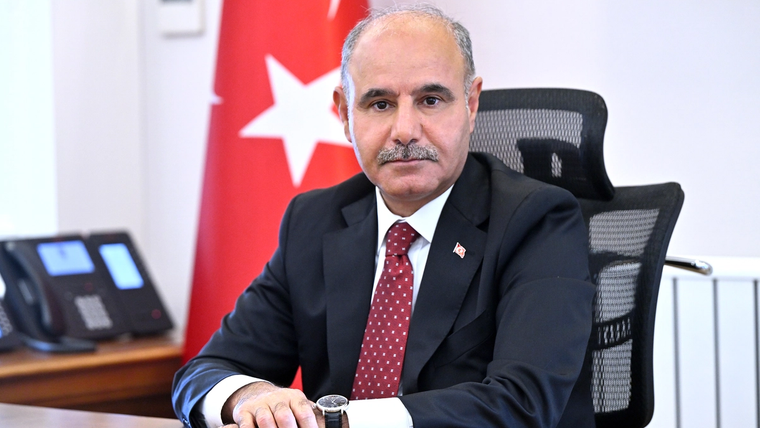 Kırmızı kravat takan bir adam, ofis ortamında oturuyor. Arkada Türk bayrağı görünmekte.