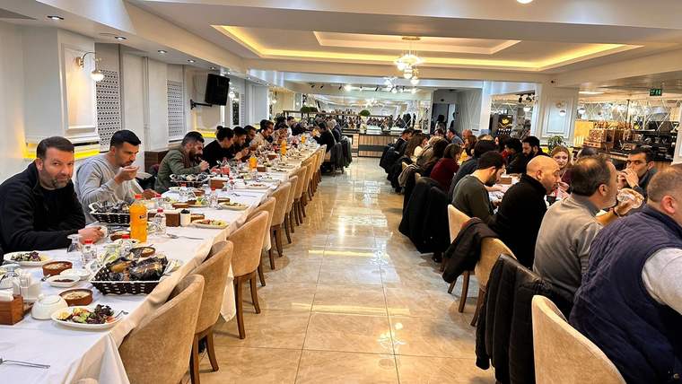 Restoran salonunda uzun masalarda oturan kalabalık grup, yemek yerken ve sohbet ederken görülüyor.