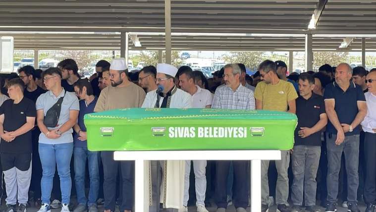 Yeşil bir tabutun önünde, kalabalık bir grup insan duruyor; bir kişi konuşma yapıyor.