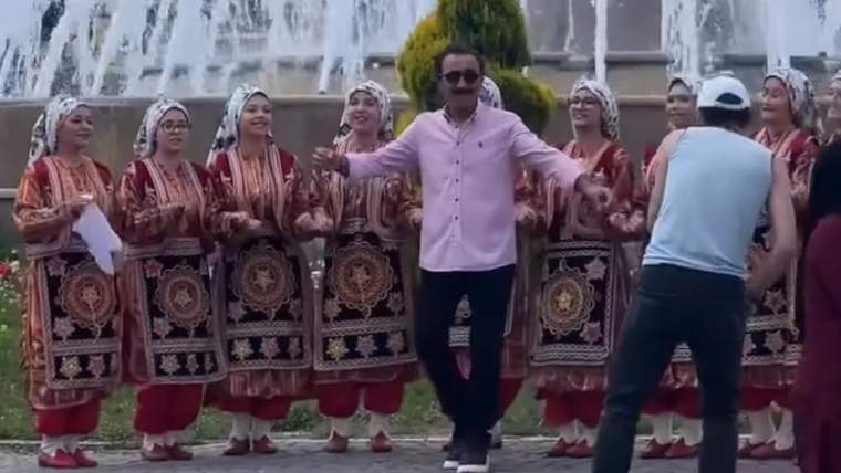 Pembe gömlekli bir kişi, çeşmeler önünde geleneksel kıyafetli kadınlarla birlikte dans ediyor.