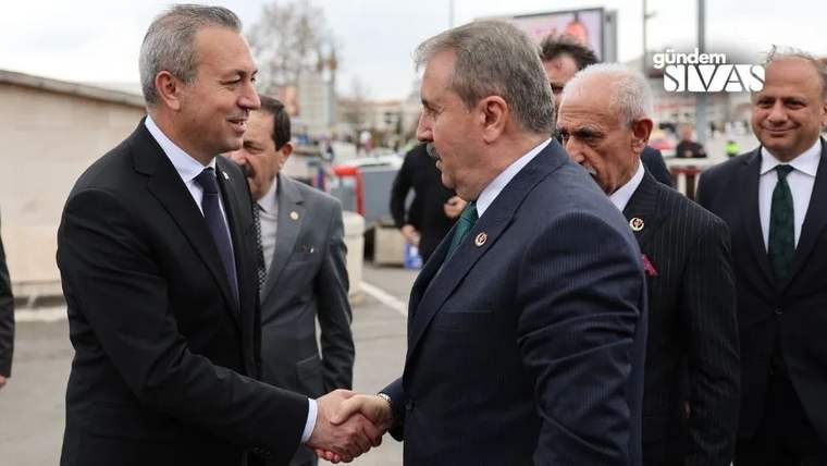 Takım elbiseli iki kişi açık havada el sıkışıyor; arkalarında takım elbiseli birkaç kişi görülüyor.