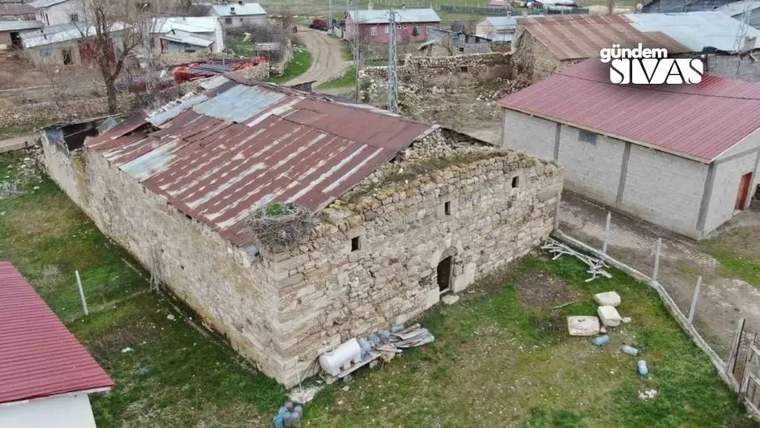 Paslı metal çatılı, taş duvarlı bir yapı; çevresinde evler, yeşil alan ve dağınık eşyalar görülüyor.