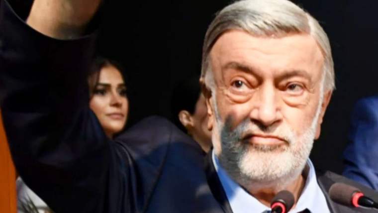 Beyaz saçlı ve sakallı bir erkek, kalabalık önünde mikrofonla konuşurken yakın planda görülüyor.