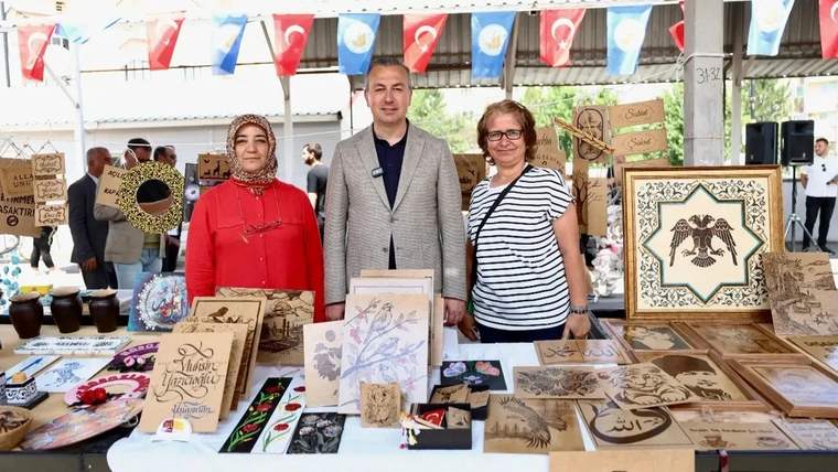 Bir stantta, el yapımı ürünlerin sergilendiği bir pazar alanında üç kişi duruyor.