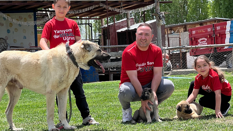 Bir adam, iki çocuk ve bir köpek, yeşil alanda oturuyor; çocuklar kucaklarında yavru köpekler tutuyor.