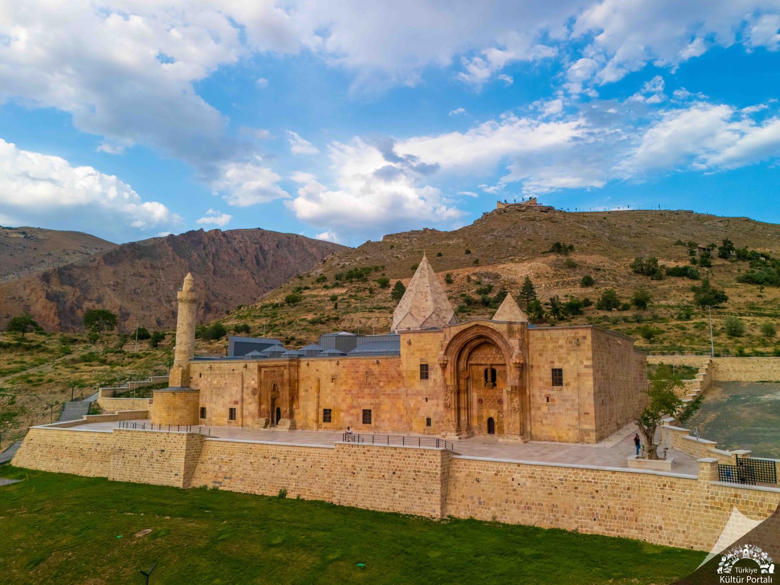 Dağlık bir arazide, taş duvarlı yapı kompleksi ve konik çatılı kuleleri görülüyor.