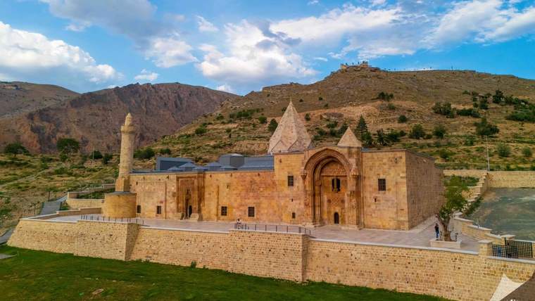 Dağlık bir arazide, taş duvarlı yapı kompleksi ve konik çatılı kuleleri görülüyor.
