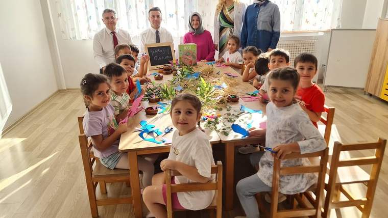Çocuklar, üzerinde el işi malzemeleri bulunan masa çevresinde otururken üç yetişkin arkalarında duruyor.