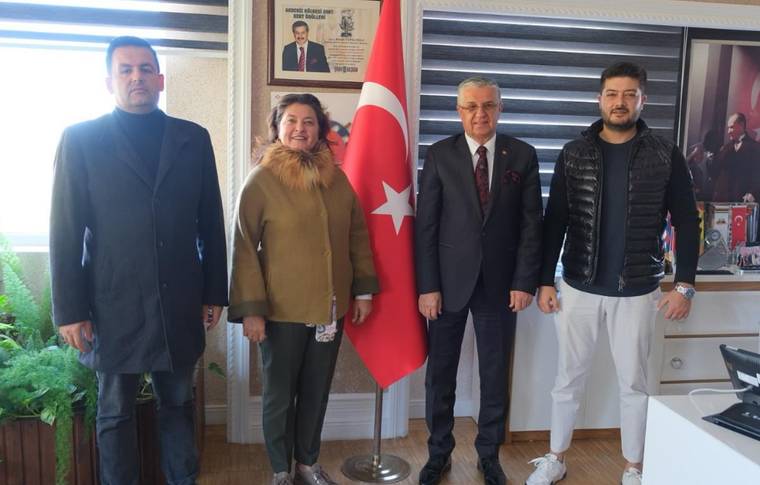 Dört kişi, Türk bayrağının önünde bir odada duruyor. Arkada çerçevelenmiş fotoğraflar var.