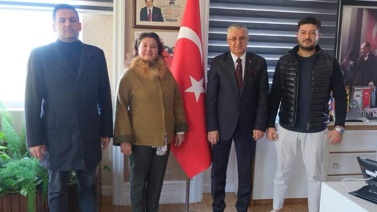 Dört kişi, Türk bayrağının önünde bir odada duruyor. Arkada çerçevelenmiş fotoğraflar var.