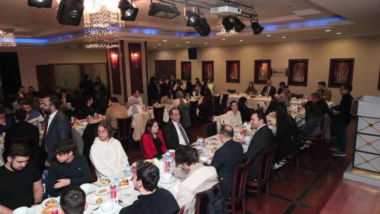 Şık bir salonda, yemek masaları çevresinde oturan ve sohbet eden kalabalık bir grup görülüyor.