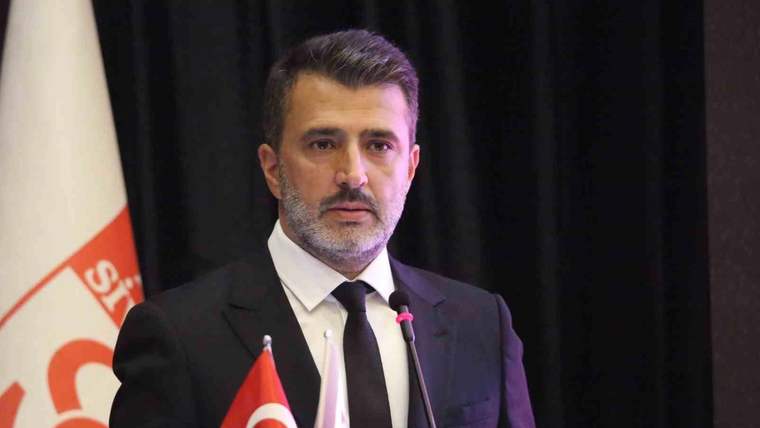Kürsüde takım elbiseli bir kişi, Türk bayrakları ve mikrofonlar önünde konuşma yapıyor.