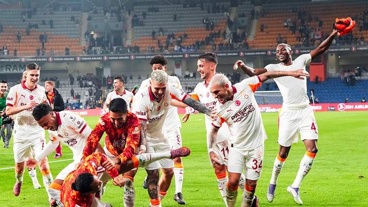 Beyaz formalı futbolcular, kırmızı formalı kalecinin yanında sahada sevinç yaşarken tribünler görülüyor.