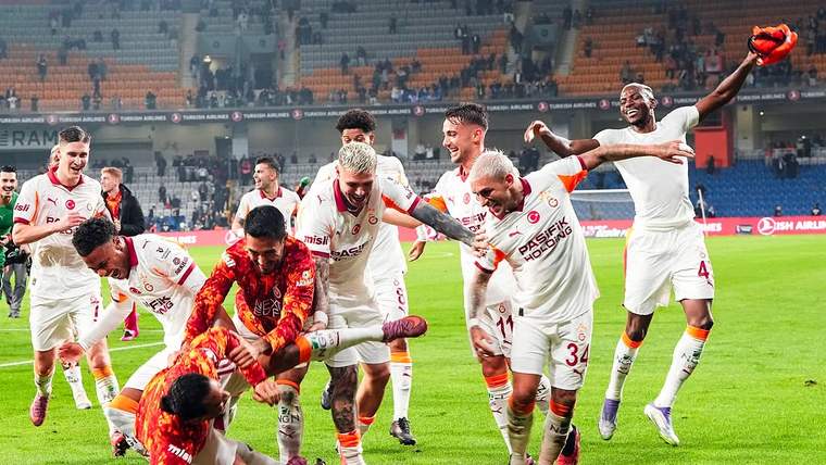 Beyaz formalı futbolcular, kırmızı formalı kalecinin yanında sahada sevinç yaşarken tribünler görülüyor.
